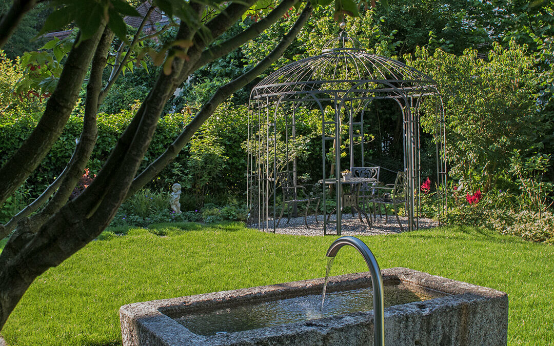Ein rechteckiger Steinbrunnen mit Wasserlauf in einem Garten mit Rasen und Bäumen.