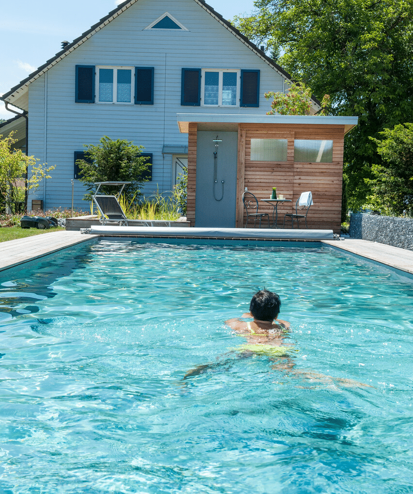 Schwimmbad mit Holzschuppen und Haus im Hintergrund Person schwimmt in einem klaren Schwimmbad vor einem Holzschuppen und einem Wohnhaus.