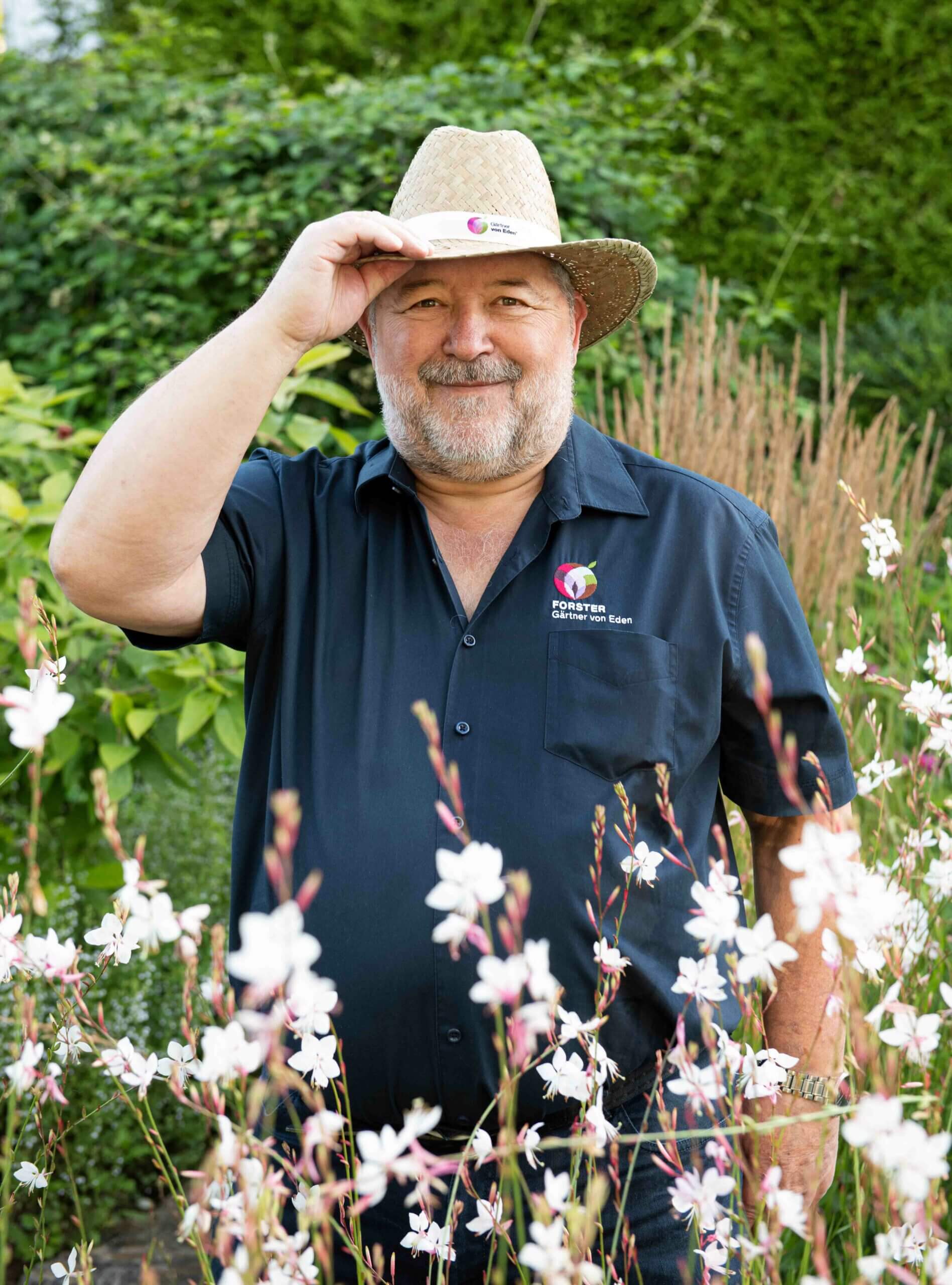 Hans-Peter Forster mit Strohhut und schwarzem Hemd steht zwischen weißen Blumen im Garten.
