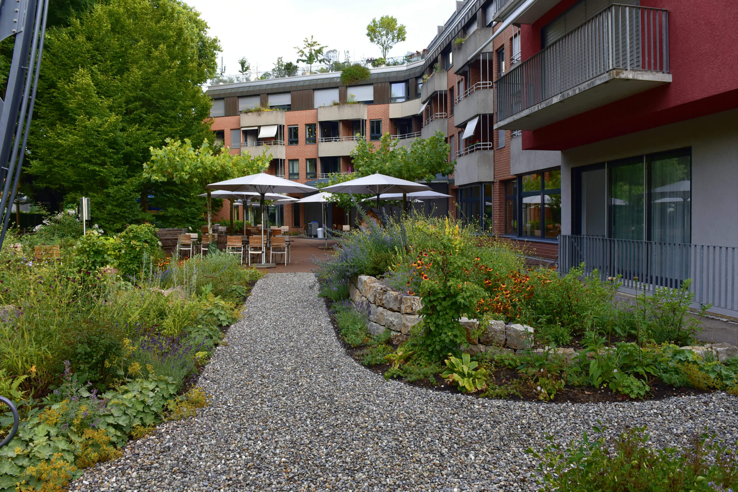 herzstueck_klein Garten mit Kieswegen, Pflanzenbeeten und Sonnenschirmen in einem Wohnbereich.