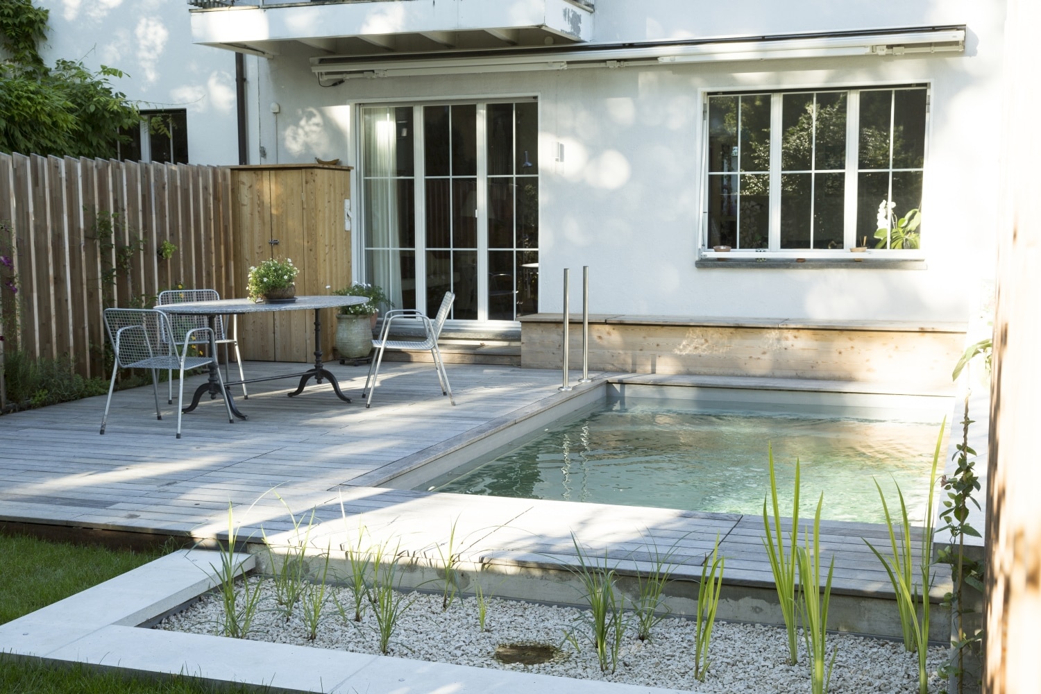 Ein kompakter Bio Minipool im kleinen Garten liegt neben einer Holzterrasse mit Tisch, Stühlen und moderner Hausfassade in ruhiger Wohnatmosphäre.