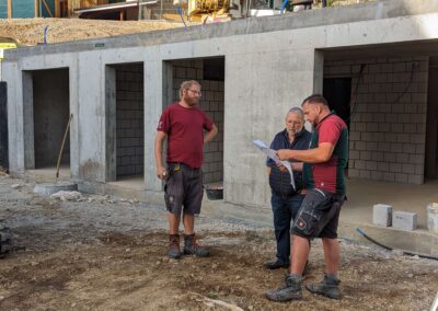 Drei Männer besprechen Baupläne vor einem Rohbau mit Betonfassade und offenen Räumen auf einer Baustelle.