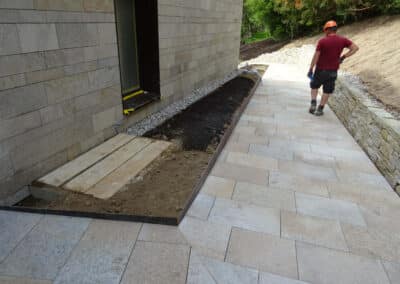 Gepflasterter Gartenweg führt an einer Natursteinmauer vorbei; ein Arbeiter geht über die neu verlegte Fläche.