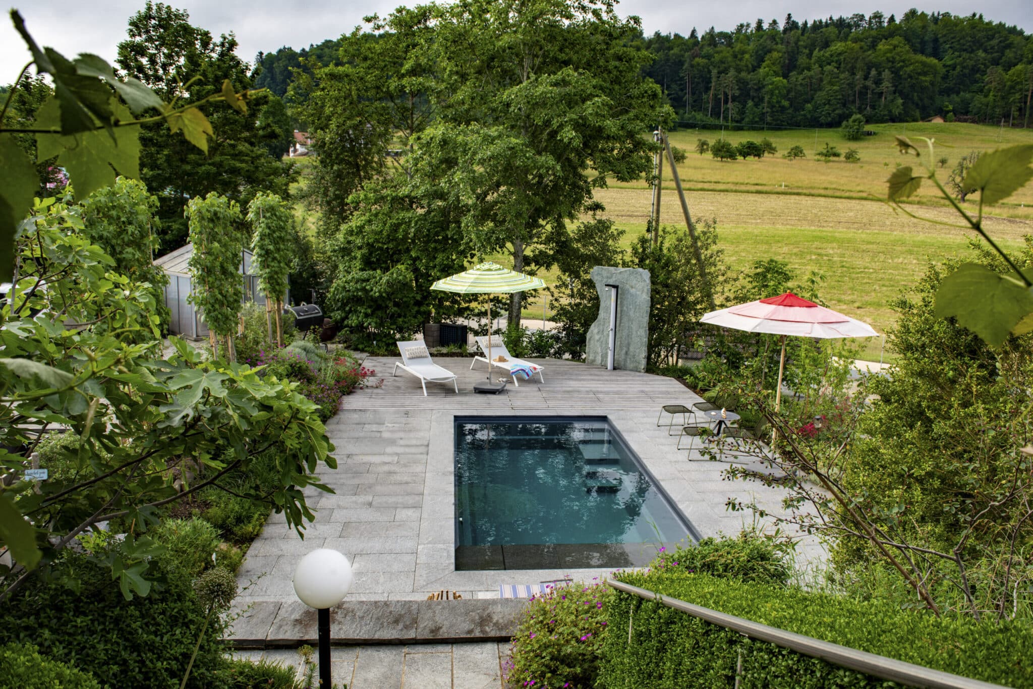 Eleganter Gartenpool auf einer Natursteinterrasse mit Sitzbereich, Sonnenschirmen und Blick über Felder und Wald