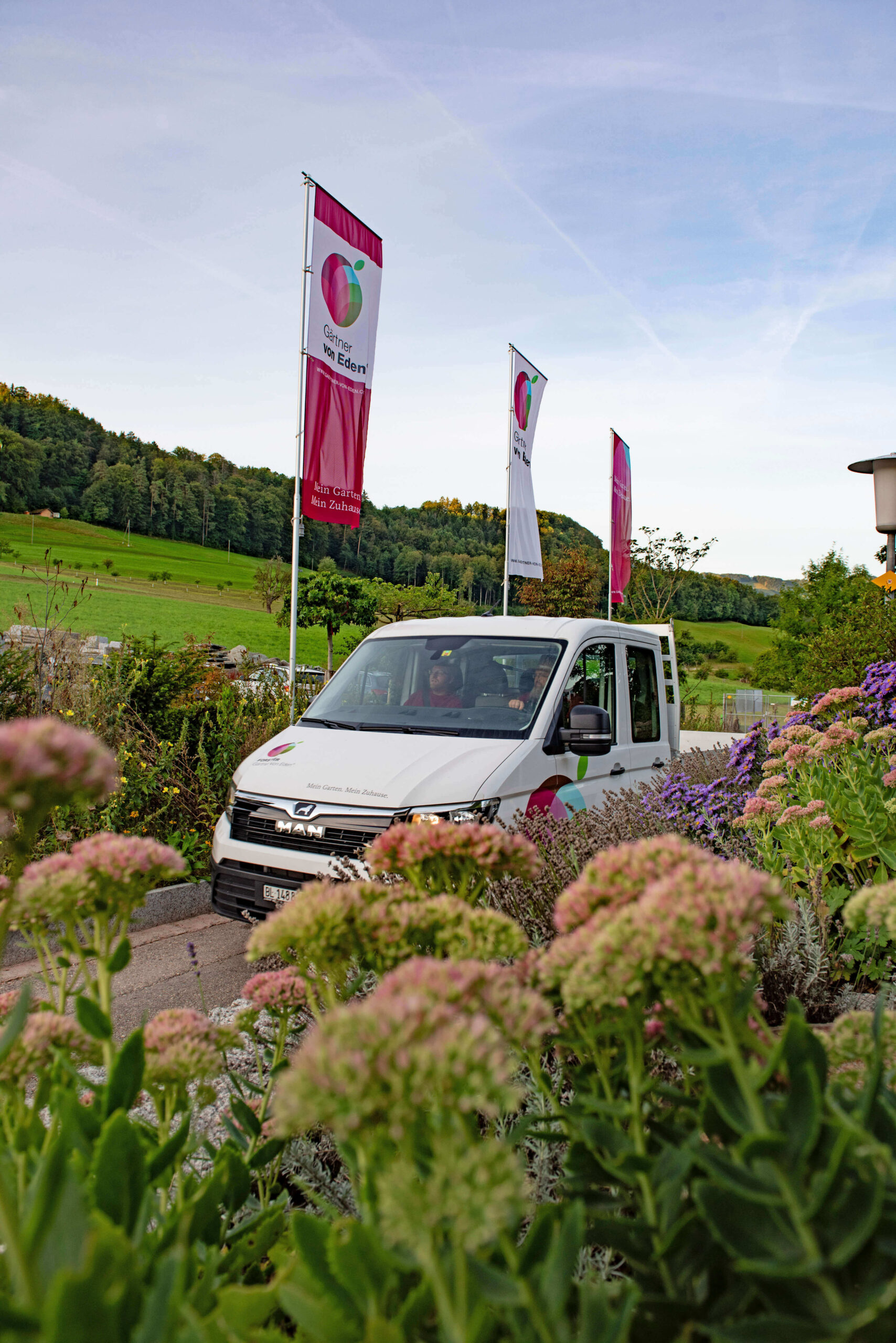 Ein weißer Gartenbau-Transporter steht zwischen blühenden Pflanzen und Fahnen von Forster Gartenbau AG vor grünen Hügeln.