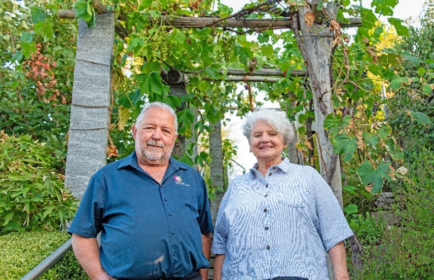 Ein älteres Paar steht unter einer begrünten Pergola im Garten und lächelt freundlich in die Kamera.