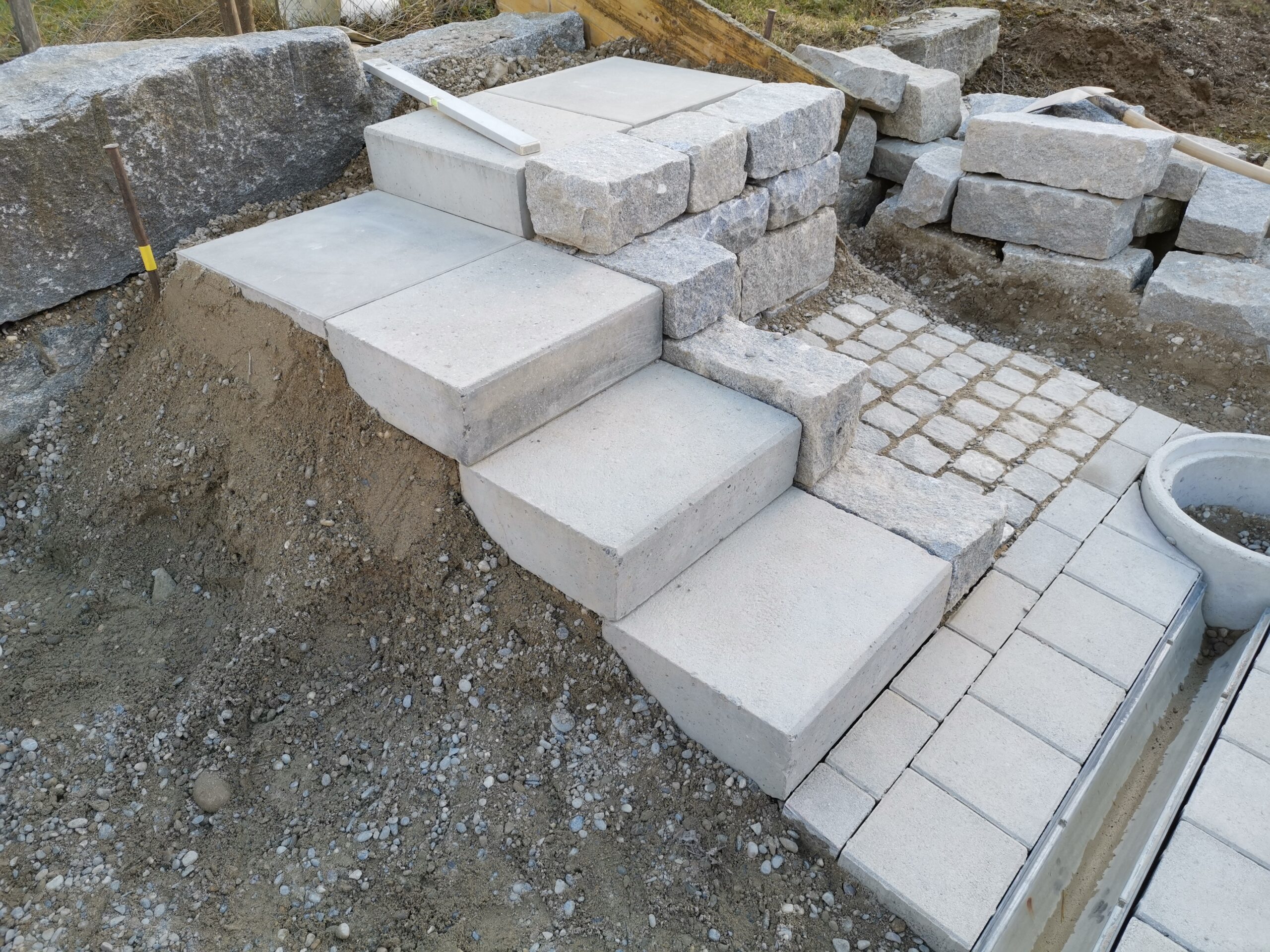 Neue Natursteintreppe aus hellen Betonplatten mit Pflasterweg und gestapelten Steinblöcken im Gartenbau