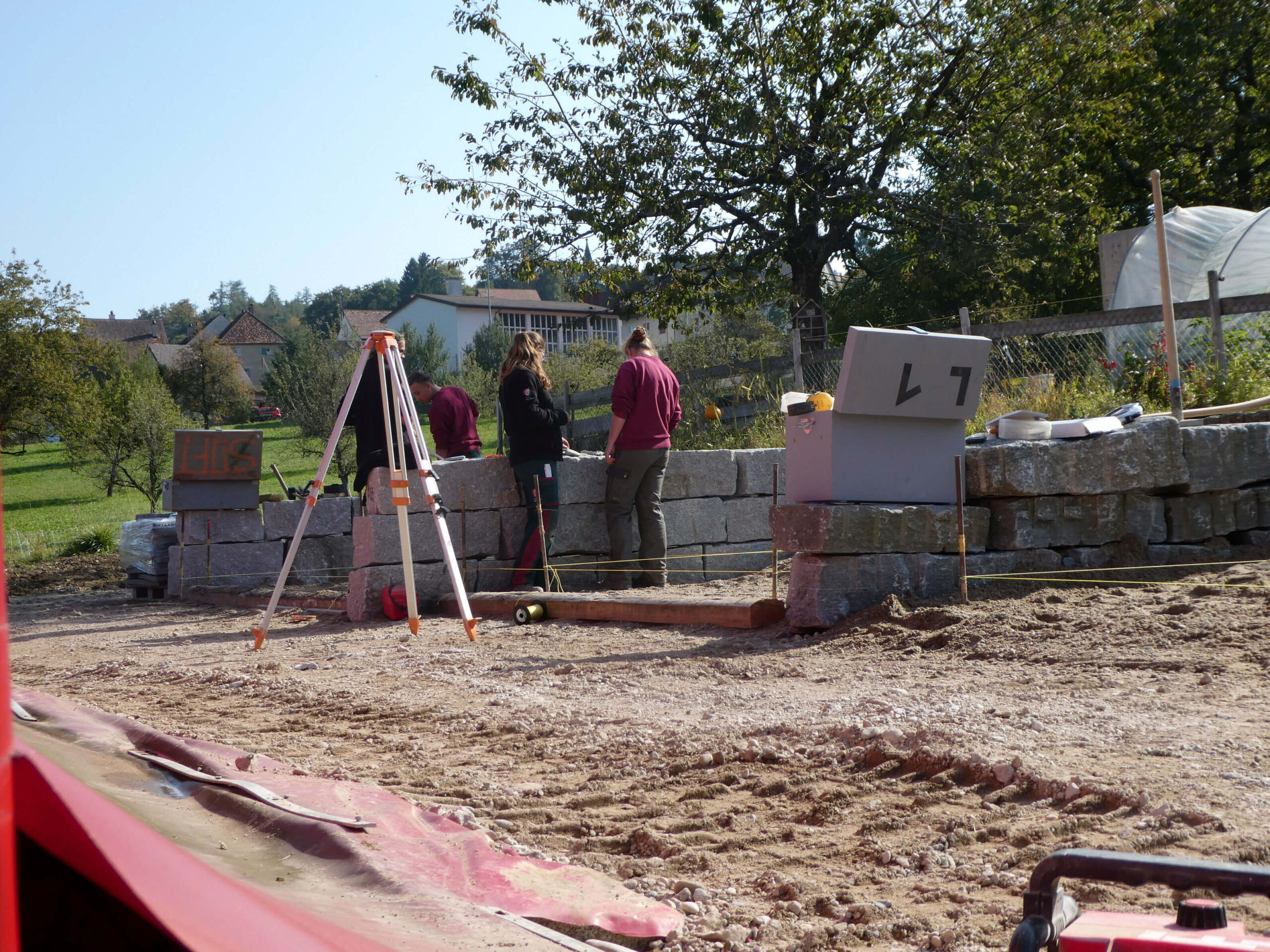 Mehrere Fachkräfte vermessen eine neue Natursteinmauer auf einer Gartenbaustelle mit Wohnhäusern und Bäumen im Hintergrund.