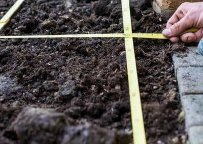 Eine Hand legt ein gelbes Maßband über dunkle Gartenerde und vermisst ein rechteckiges Bodenstück neben Pflastersteinen.