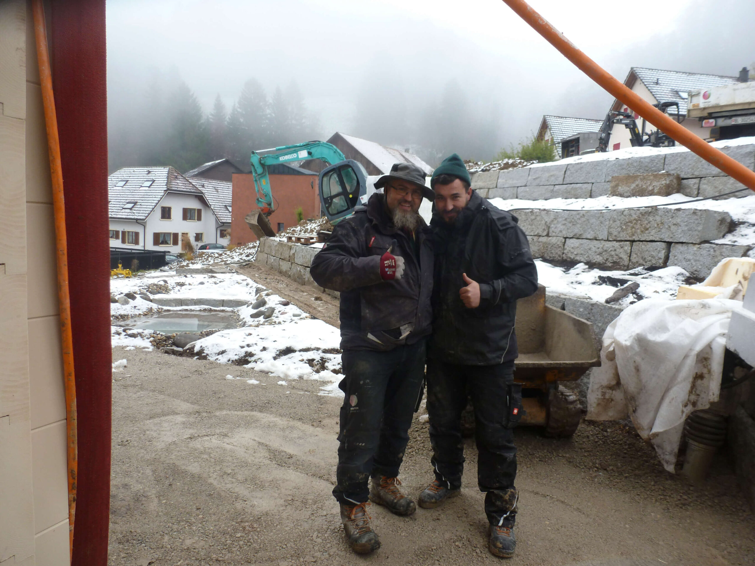 Zwei Gartenbauer stehen bei einer verschneiten Baustelle neben Minibagger, Transportkarre und Natursteinmauer im Bergdorf.